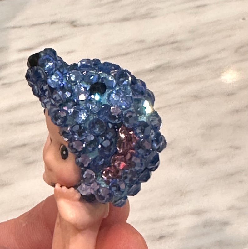 Puede incluir: Una cabeza de mu&ntilde;eca vintage con un adorno para el pelo con joyas azules. La cara de la mu&ntilde;eca es p&aacute;lida con rasgos delicados, y el adorno para el pelo es un grupo de gemas azules. La mu&ntilde;eca es sostenida por una mano.