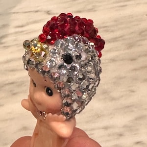 Puede incluir: Una peque&ntilde;a figura con rostro y cuerpo rosados, adornada con un tocado de joyas plateadas, rojas y amarillas brillantes. La figura tiene ojos negros y est&aacute; sujeta por los dedos de una persona.