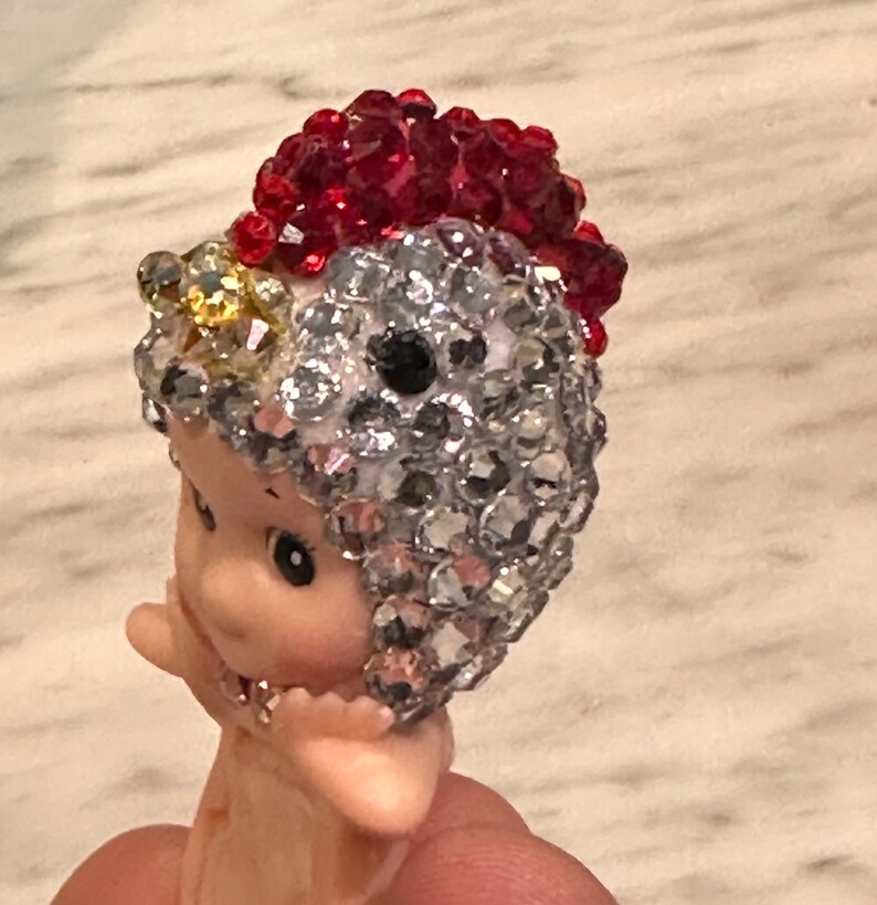 Puede incluir: Una peque&ntilde;a figura con rostro y cuerpo rosados, adornada con un tocado de joyas plateadas, rojas y amarillas brillantes. La figura tiene ojos negros y est&aacute; sujeta por los dedos de una persona.