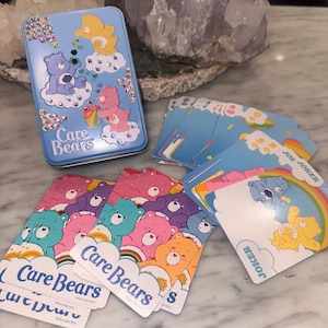 Peut inclure: Une boîte en métal bleu clair Care Bears décorée de strass, accompagnée d'un jeu de cartes représentant des personnages Care Bears, des arcs-en-ciel et des nuages. Les cartes affichent le logo Care Bears et le mot Joker sur certaines cartes.