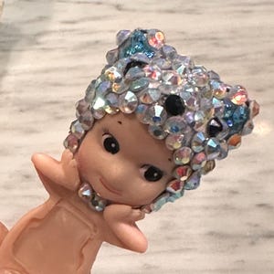 Puede incluir: Peque&ntilde;a figura de color rosa claro de una figura querub&iacute;nica con un tocado enjoyado. El tocado est&aacute; adornado con cristales iridiscentes, transparentes y azules. La figura tiene una expresi&oacute;n serena y se sostiene las manos cerca de la cara.
