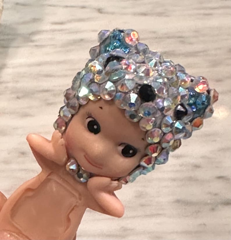 Puede incluir: Peque&ntilde;a figura de color rosa claro de una figura querub&iacute;nica con un tocado enjoyado. El tocado est&aacute; adornado con cristales iridiscentes, transparentes y azules. La figura tiene una expresi&oacute;n serena y se sostiene las manos cerca de la cara.