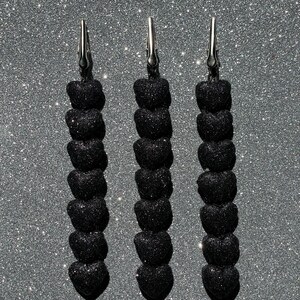 Könnte beinhalten: Drei vertikale Roach-Clips, jeweils mit einer silbernen Metall-Krokodilklemme an der Oberseite, die an einer Schnur aus sieben kleinen, schwarzen, glitzernden herzförmigen Perlen befestigt ist. Die Herzen sind in einer Reihe angeordnet.