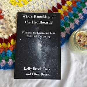 May include: A book titled "Who's Knocking on the Headboard?" with the subtitle "Guidance for Embracing Your Spiritual Awakening" is displayed. The book is black with white text and a galaxy image. A lit candle in a glass jar is next to the book.