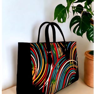 May include: Black tote bag with a vibrant, colorful design. The bag features a pattern of curved lines in red, yellow, blue, and white against a black background. It has black handles and a rectangular shape, suitable for carrying items.