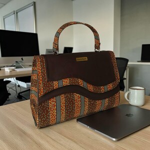 Puede incluir: Un bolso marrón con un diseño de tela estampada a rayas verticales naranjas, rojas y azules. El bolso tiene una solapa y un asa de color marrón oscuro, con el texto "Love & Wealth" en una pequeña placa dorada.