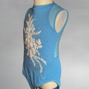 May include: A light blue leotard with a high neckline and a decorative floral design on the front. The leotard is embellished with small, sparkling rhinestones and features sheer mesh panels on the sides and shoulders.