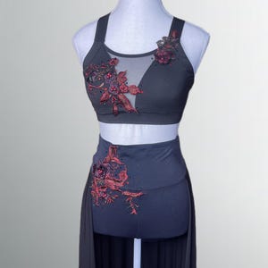 Puede incluir: Un conjunto de baile de dos piezas gris oscuro sobre un maniquí. La parte superior es un sujetador deportivo con un panel de malla transparente y bordados florales rojos. La parte inferior de cintura alta tiene un diseño floral a juego y una falda larga y fluida.