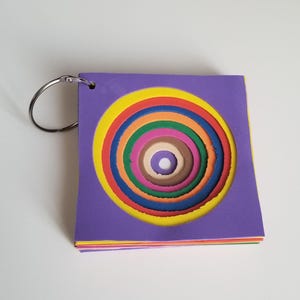 May include: A stack of colorful paper circles in a spiral design, bound together with a silver ring. The top sheet is purple with a yellow circle, followed by red, orange, green, pink, brown, and white. The edges of the paper are visible.