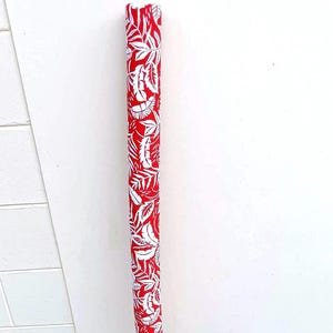 May include: A rolled-up fabric with a red and white pattern. The fabric displays a repeating design of white tropical leaves on a bright red background. The roll is cylindrical and seems to be made of a flexible material.