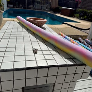 May include: A rolled yoga mat with a tie-dye design in pink, yellow, and blue is on a white tiled surface. A small, grey object is nearby. Other rolled mats are visible in the background near a swimming pool.