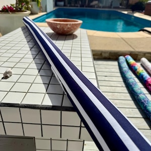 May include: A long, rolled navy blue and white striped fabric roll rests on a white tiled surface. The fabric has a nautical design. Other patterned rolls are visible in the background, along with a pool and outdoor setting.