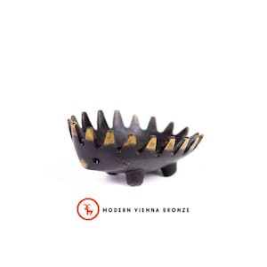 Op de afbeelding: Een kleine, zwarte bronzen kom in de vorm van een egel met gouden accenten op de stekels. De kom heeft drie kleine pootjes en staat tegen een witte achtergrond. De tekst "MODERN VIENNA BRONZE" staat eronder.
