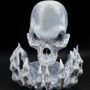May include: A silver-colored skull sculpture with a detailed, textured surface. The skull is held by two hands with sharp, claw-like fingers. The sculpture is set against a black background, highlighting the intricate details of the design.