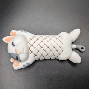 Puede incluir: Una figura con forma de gato blanco y gris, tumbada boca arriba. El gato tiene un patrón texturizado en forma de escamas en la espalda y un pequeño lazo gris en el extremo. La cara del gato tiene detalles grises y naranjas.
