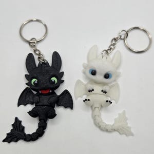 May include: Two dragon keyrings. One is black with green eyes and a red mouth, the other is white with blue eyes. Both have wings and tails, and are attached to silver keyrings.