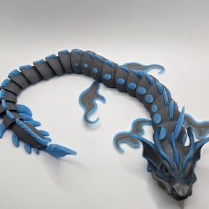 May include: A gray and blue articulated dragon figurine. The dragon has a segmented body with blue accents on its fins and head. The figurine is posed on a white surface.