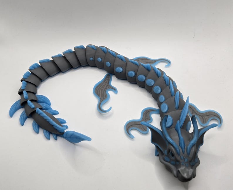 May include: A gray and blue articulated dragon figurine. The dragon has a segmented body with blue accents on its fins and head. The figurine is posed on a white surface.