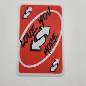 May include: A red and white card with the words "LOVE YOU MORE" and a reverse arrow symbol. The card is a novelty item, likely a custom version of a popular card game.