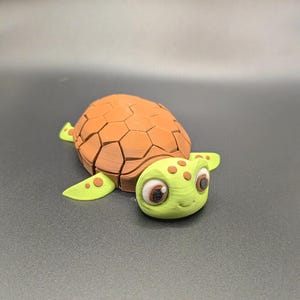 May include: A 3D-printed turtle figurine with a brown, segmented shell and a light green head and flippers. The turtle has large, expressive eyes with brown irises and small, brown spots on its head and flippers. The figurine is on a dark gray surface.