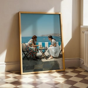 May include: A framed print shows two people playing chess outdoors. They are seated at a small table with a chessboard, overlooking a blue sea and sky. The print is in a wooden frame, leaning against a wall.