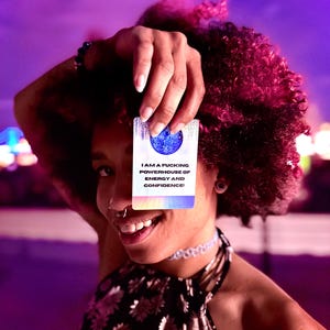 May include: A rectangular affirmation card featuring a blue disco ball graphic at the top and the text I AM A FUCKING POWERHOUSE OF ENERGY AND CONFIDENCE! printed in black, with a colorful grid pattern at the bottom edge against a white background.