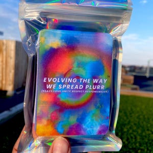 May include: A rectangular deck of cards with a vibrant tie-dye pattern in shades of blue, yellow, and red, enclosed in a holographic resealable plastic pouch. The text reads: EVOLVING THE WAY WE SPREAD PLURR (PEACE, LOVE, UNITY, RESPECT, RESPONSIBILITY).