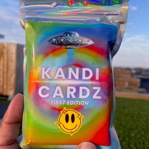 May include: A holographic, iridescent pouch containing a deck of cards. The front features a graphic of a UFO, a rainbow-colored circular background, and the text KANDI CARDZ FIRST EDITION. A yellow smiley face icon is printed at the bottom.