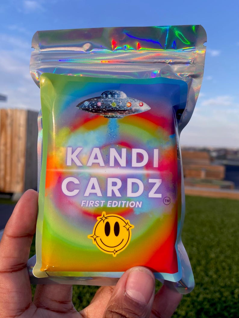 May include: A holographic, iridescent pouch containing a deck of cards. The front features a graphic of a UFO, a rainbow-colored circular background, and the text KANDI CARDZ FIRST EDITION. A yellow smiley face icon is printed at the bottom.