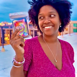 May include: A rectangular deck of cards enclosed in a clear plastic pouch featuring a vibrant, multi-colored tie-dye pattern with swirling hues of pink, yellow, orange, and blue, displaying small white text on the front surface of the card deck.