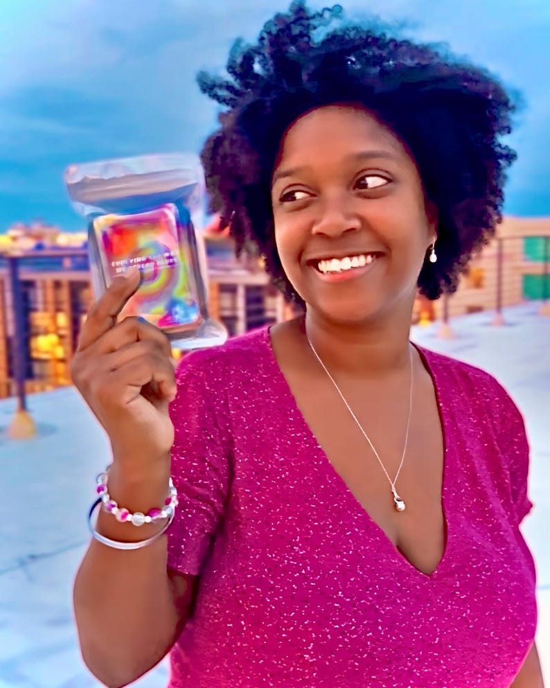 May include: A rectangular deck of cards enclosed in a clear plastic pouch featuring a vibrant, multi-colored tie-dye pattern with swirling hues of pink, yellow, orange, and blue, displaying small white text on the front surface of the card deck.