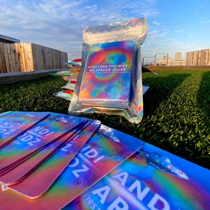 May include: A stack of rectangular cards with a holographic rainbow finish. The cards feature the text "ANDI ARDZ FIRST EDITION" and a yellow smiley face. One pack is sealed in a clear holographic pouch with the text "EVOLVING THE WAY WE SPREAD PLURR".