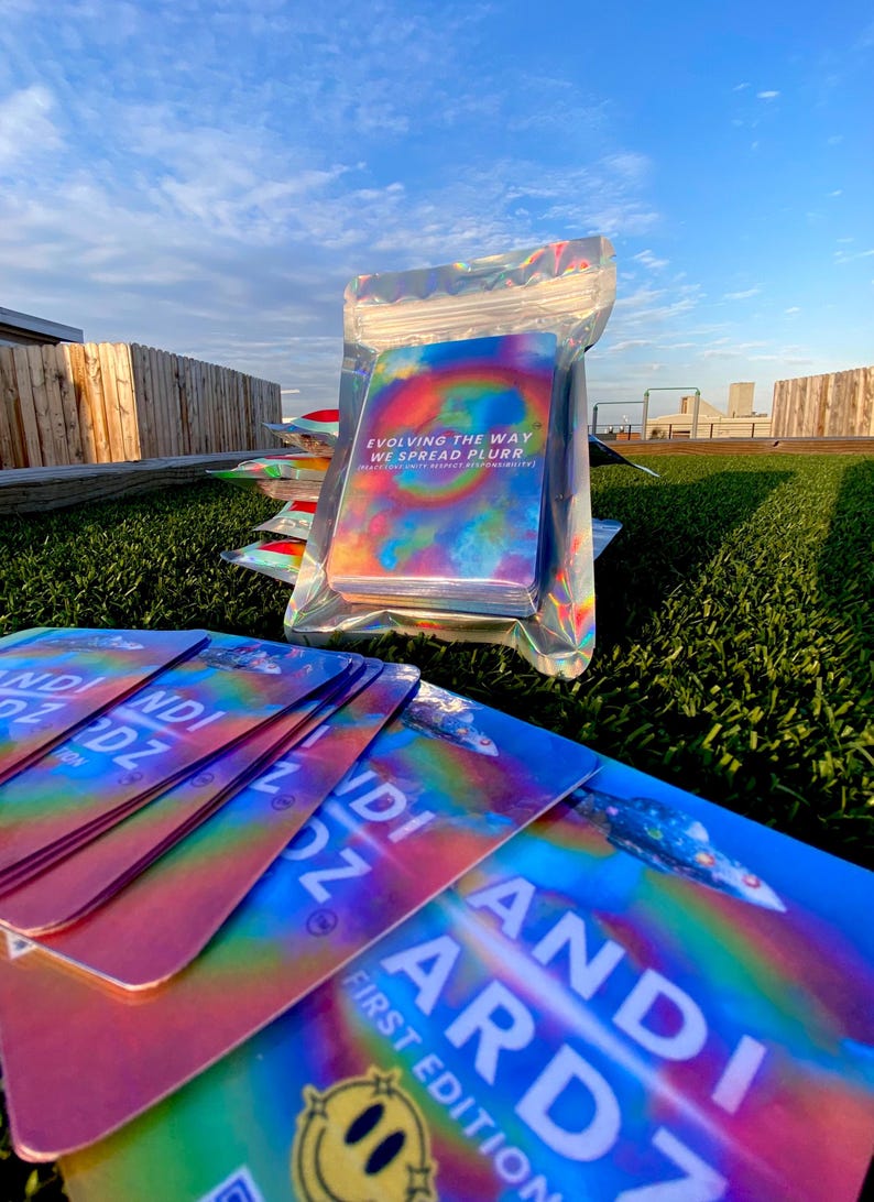 May include: A stack of rectangular cards with a holographic rainbow finish. The cards feature the text "ANDI ARDZ FIRST EDITION" and a yellow smiley face. One pack is sealed in a clear holographic pouch with the text "EVOLVING THE WAY WE SPREAD PLURR".