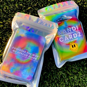 May include: Two holographic, iridescent resealable pouches containing card decks. One pouch displays a UFO graphic, the text KANDI CARDZ FIRST EDITION, and a smiley face. The other shows the text EVOLVING THE WAY WE SPREAD PLURR.