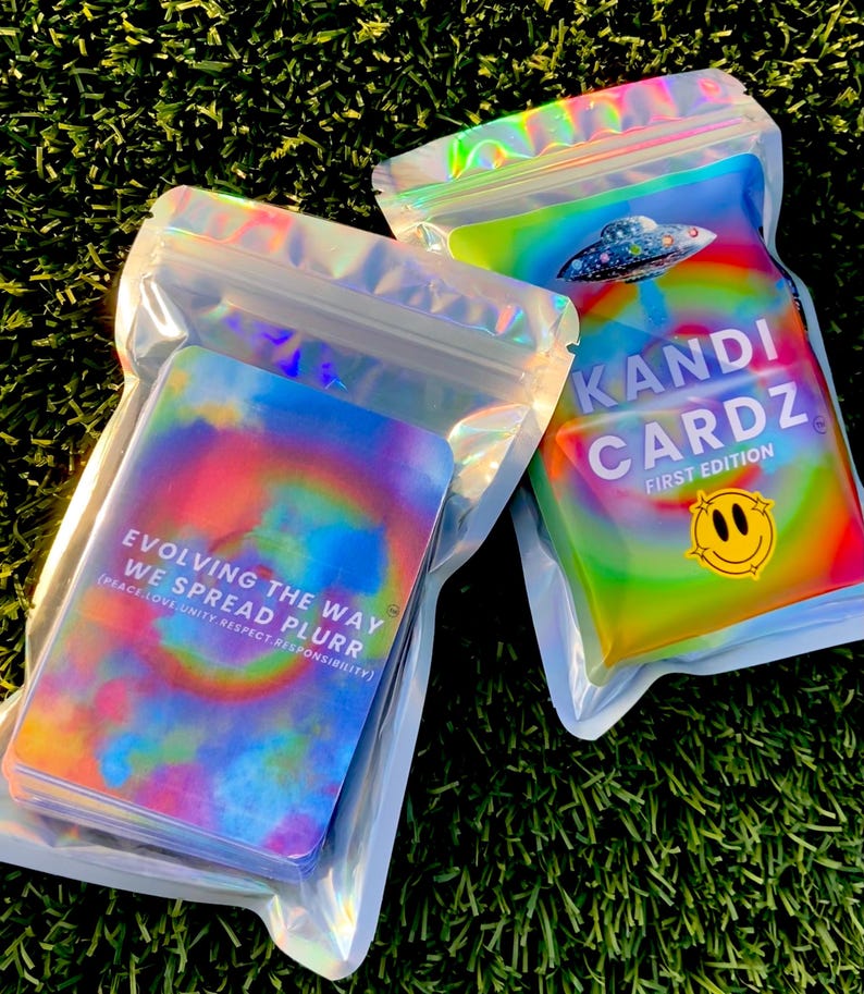 May include: Two holographic, iridescent resealable pouches containing card decks. One pouch displays a UFO graphic, the text KANDI CARDZ FIRST EDITION, and a smiley face. The other shows the text EVOLVING THE WAY WE SPREAD PLURR.