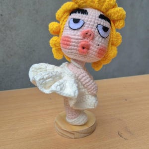 May include: Crocheted doll with yellow hair and a white dress, standing on a wooden base. The doll has a surprised expression with pursed lips and rosy cheeks. The doll is handmade and appears to be a unique art piece.