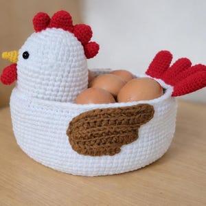 Op de afbeelding: Een witte gehaakte kippenvormige mand gevuld met bruine eieren. De kip heeft een rode kam en lellen, een gele snavel en een bruin vleugeldetail. De tekst "AmigurumibyroseStore" staat onderaan.