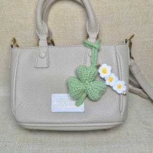 May include: A beige pebbled leather handbag with two top handles and a detachable shoulder strap. A white rectangular label with the numbers 0 through 23 is stitched to the front. A green crocheted four-leaf clover and three small white flowers are attached.