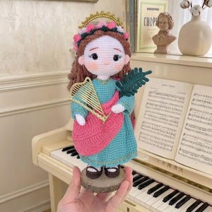 May include: A handcrafted crochet doll with brown hair, a pink floral crown, and a gold halo. The doll wears a teal dress with a pink sash, holding a gold wire harp and a green palm leaf. It stands on a circular wooden base, featuring dark shoes.