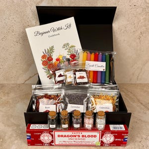 May include: A black gift box containing a Beginner Witch Kit guidebook, a set of multi-coloured spell candles, various dried herbs including rose, lavender, and calendula in clear bags, five small glass jars with cork stoppers, and incense.