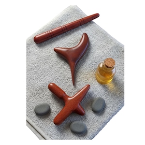 Könnte beinhalten: Eine Sammlung von Massage-Tools und Zubehör. Das Bild zeigt mehrere Massage-Tools aus Holz in verschiedenen Formen, darunter ein Stab, ein Y-förmiges Werkzeug und ein kreuzförmiges Werkzeug. Eine kleine Glasflasche mit Korken und einige graue Steine sind ebenfalls vorhanden.