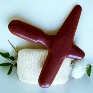 Può includere: Un attrezzo da massaggio in legno rosso scuro con una testa a forma di croce e un manico. L'attrezzo è appoggiato su un quadrato bianco strutturato. Lo sfondo include vegetazione e un fiore bianco.