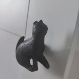 Peut inclure: Un crochet en fonte gris foncé en forme de chat. Le chat est en position assise, la queue enroulée et la tête levée. Le crochet est monté sur une surface blanche.