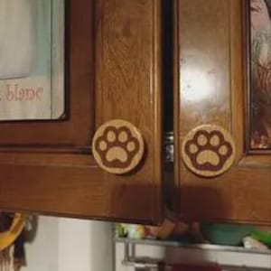 Puede incluir: Pomos de armario de madera con diseño de huella de pata. Los pomos son redondos, con un fondo marrón claro y una huella de pata marrón oscuro. Fijados a un armario de madera.