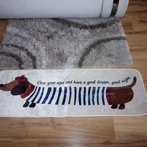 Könnte beinhalten: Rechteckiger, cremefarbener Teppich mit einem Cartoon-Dackel mit Baskenmütze und gestreiftem Hemd. Der Text "Close your eyes and have a good dream, good night" ist auf dem Teppich aufgedruckt. Ein grauer Teppich ist im Hintergrund.
