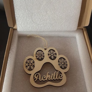 Peut inclure: Ornement en bois en forme d'empreinte de patte avec le nom "Achille" découpé. L'empreinte de patte comporte des motifs de flocons de neige dans les coussinets. L'ornement est dans une boîte en carton avec un rembourrage blanc.