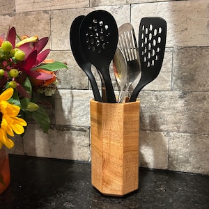 May include: A wooden utensil holder with a hexagonal shape holds several black cooking utensils and a silver spatula. A bouquet of flowers with sunflowers, orange lilies, and other colorful blooms sits to the left of the holder.