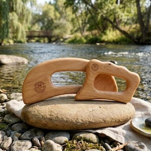 May include: Two light brown wooden massage tools with ergonomic handles and smooth, curved edges. Each tool features a circular brand logo stamped on the surface. The tools are positioned together, showcasing their distinct shapes and natural wood grain.
