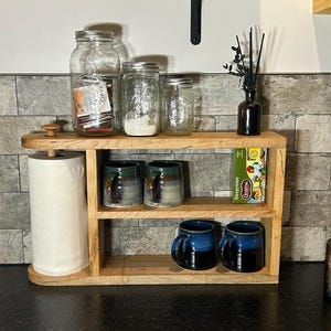May include: A wooden kitchen shelf with a paper towel holder, jars, mugs, and a box of Sleepytime tea. The shelf has two levels and is made of light-colored wood. The mugs are blue and green.