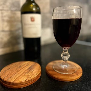 May include: A glass of red wine on a wooden coaster. A stack of wooden coasters is in the foreground. A wine bottle is in the background. The coasters and the base of the wine glass are oval-shaped.